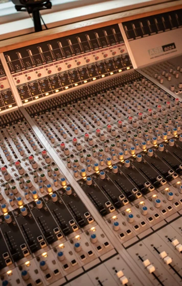 A wide shot of a professional recording studio mixing desk, with multiple sliders, knobs, and glowing lights. A wide shot of a professional recording studio mixing desk, with multiple sliders, knobs, and glowing lights.
